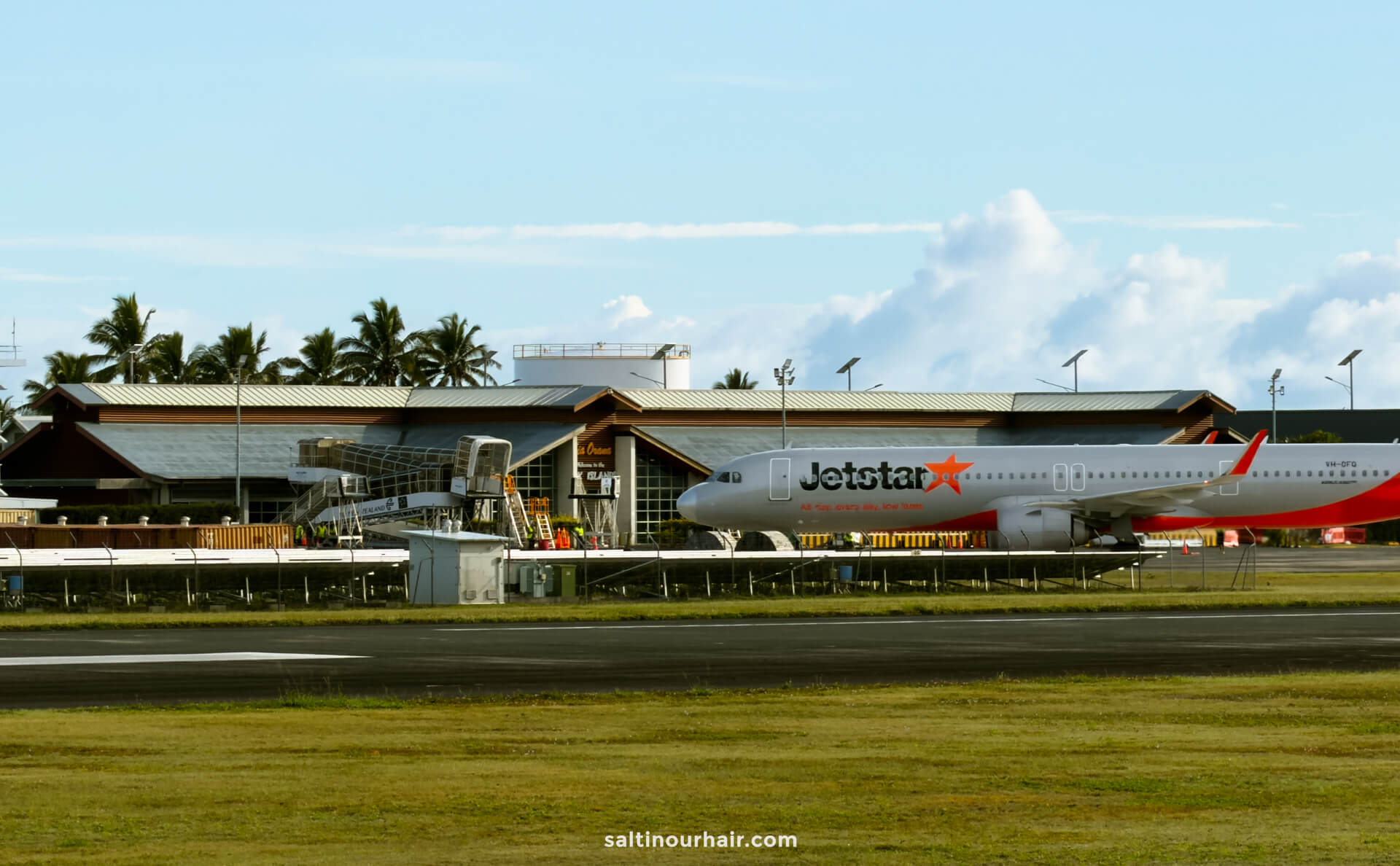 A Jetstar airplane is parked at an airport gate, with the terminal building and palm trees visible in the background&mdash;reminding travelers of exciting things to do in Rarotonga just beyond the terminal.