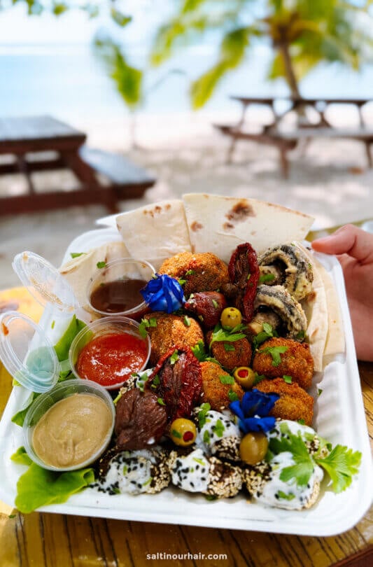 A takeout box with pita bread, falafel, dips, stuffed grape leaves, olives, sun-dried tomatoes, and garnishes&mdash;perfect for a picnic by the beach in Rarotonga. Enjoy local flavors while exploring fun things to do Rarotonga has to offer.