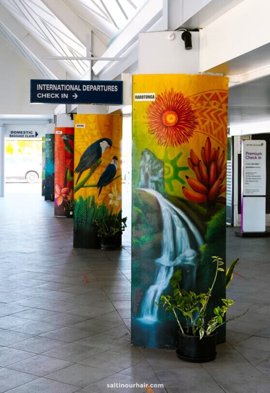 Airport interior with painted columns featuring birds and waterfalls, directional signs for international and domestic departures, and potted plants at the base&mdash;setting the scene for your arrival before exploring all the things to do in Rarotonga.
