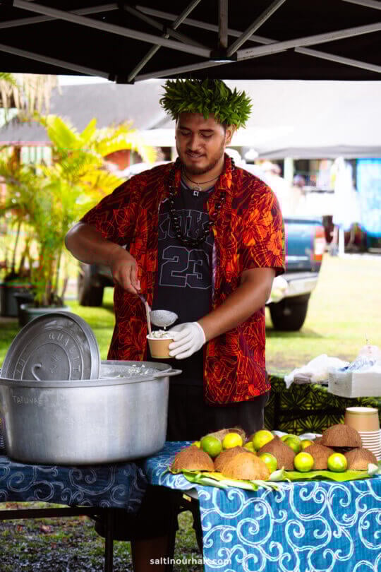 A person wearing a leafy headpiece and gloves serves food from a large pot into a cup at an outdoor market stall&mdash;one of the unique things to do in Rarotonga, with fresh breadfruit displayed on the table.