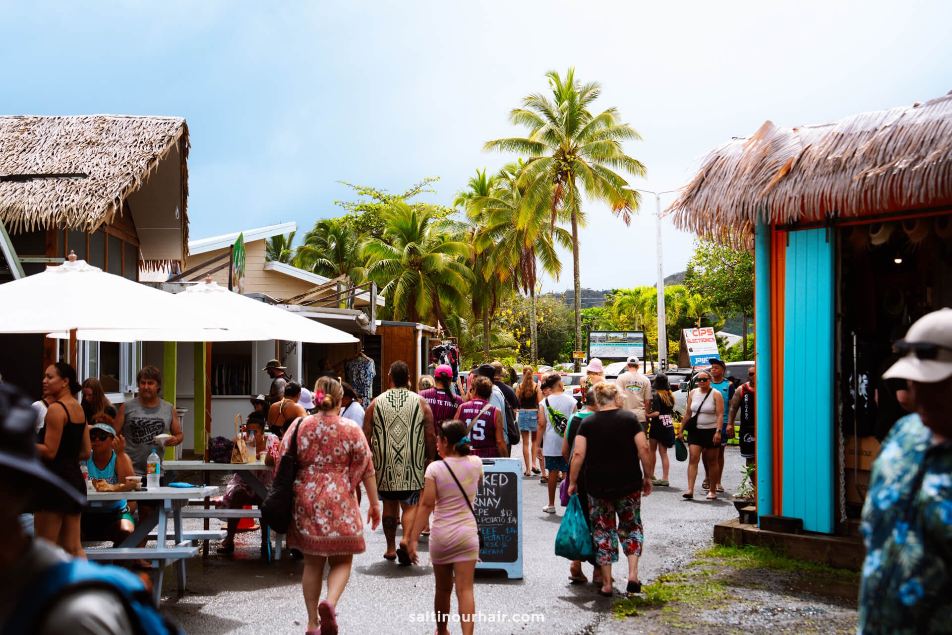 A busy outdoor market scene with people walking, shopping, and sitting under umbrellas, surrounded by tropical palm trees and colorful buildings&mdash;one of the vibrant things to do in Rarotonga, Cook Islands