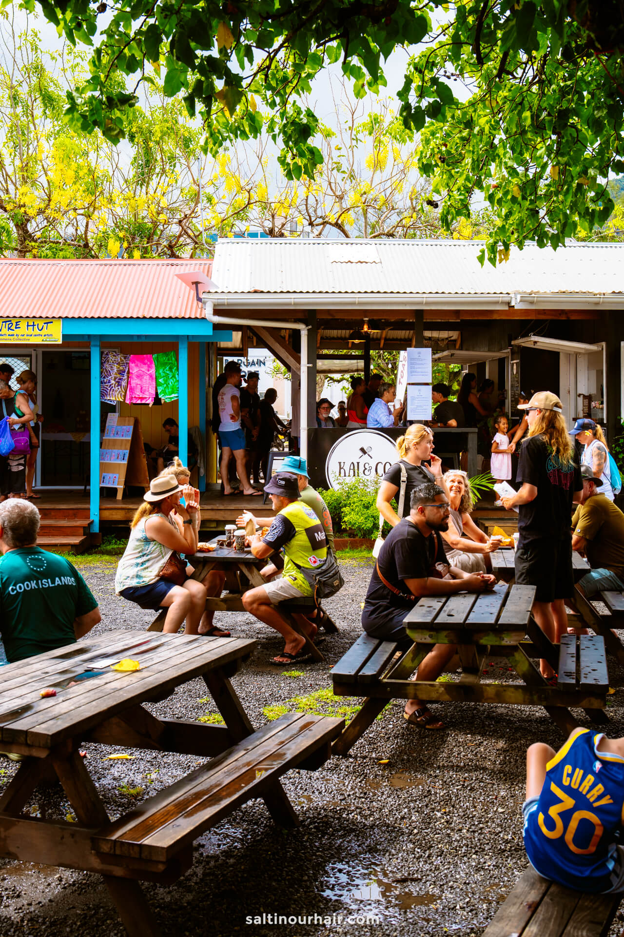 People sit at wooden picnic tables eating and chatting in an outdoor food market area, surrounded by colorful huts and menu boards&mdash;a lively spot that belongs on any things to do Rarotonga list.