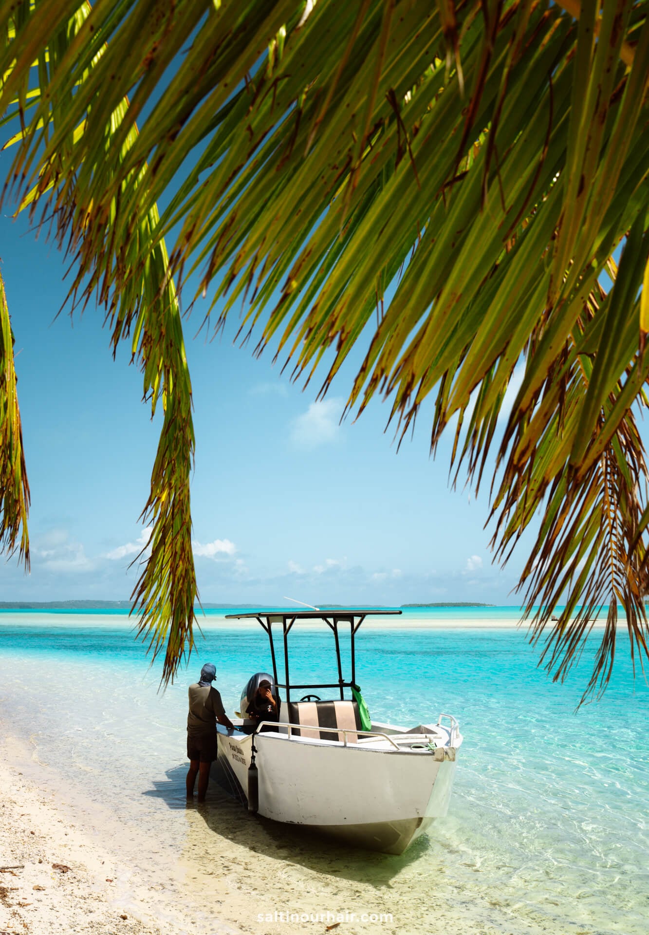 A small boat is anchored in shallow turquoise water near the sandy beach of Aitutaki island, with two people standing on the shore and palm fronds hanging overhead.