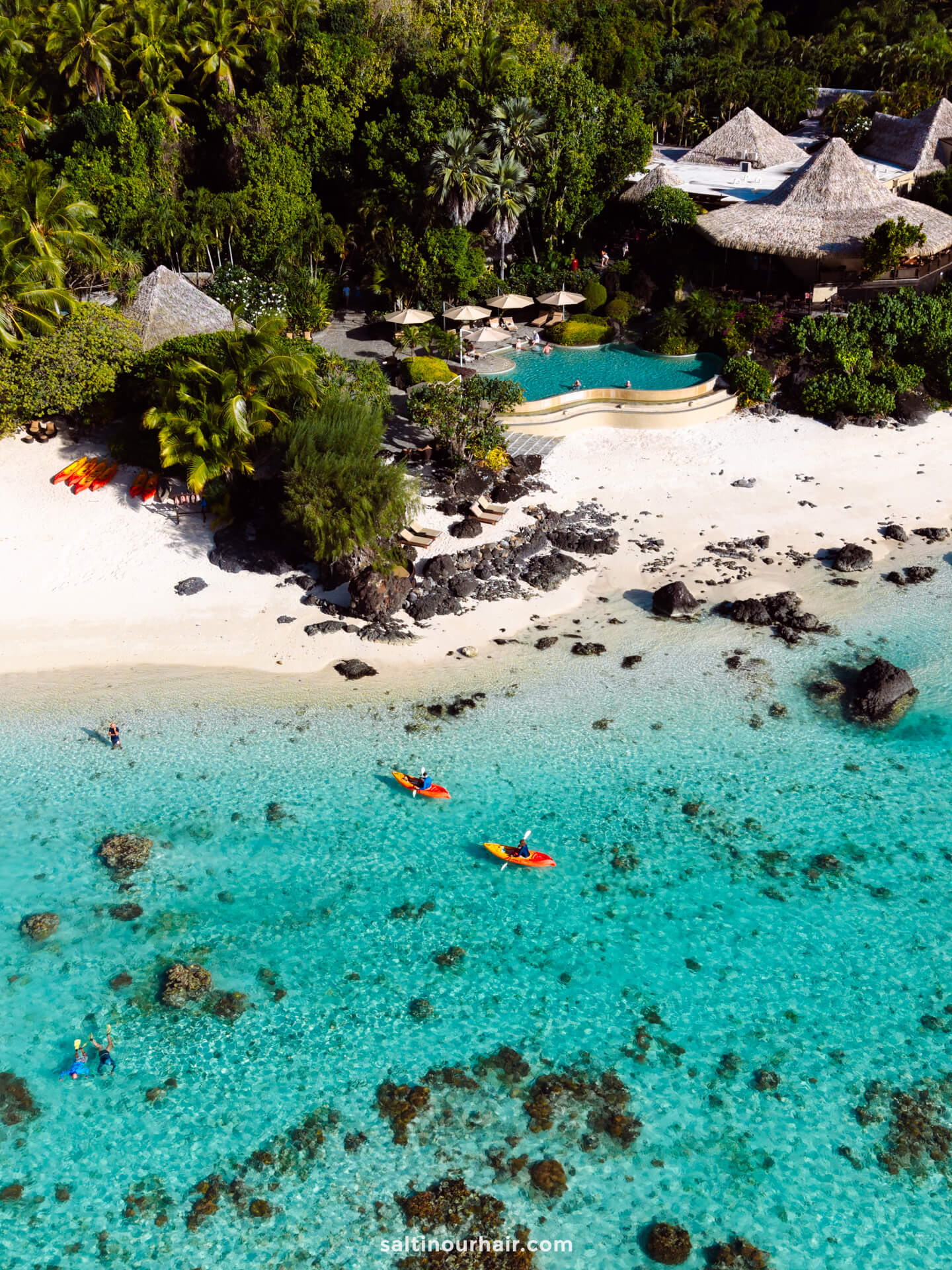 Aerial view of a tropical Aitutaki island resort with thatched-roof huts, a pool, sandy beach, and people kayaking in clear turquoise water.