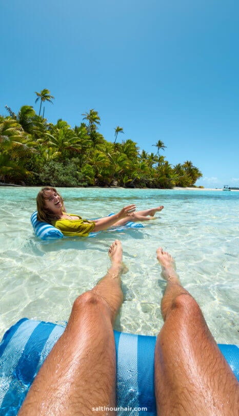 A person on a blue float relaxes in the shallow, clear tropical waters of Aitutaki island near palm trees; another person