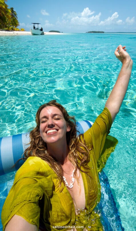 Woman in a green shirt smiles and takes a selfie while floating on a pool lounger in clear, turquoise water near the tropical beach of Aitutaki island, cook islands
