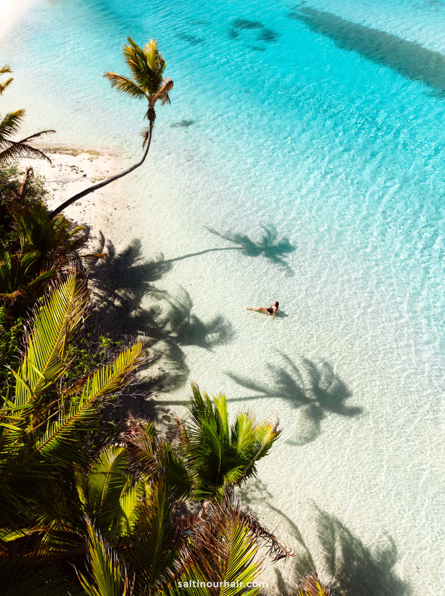 A person floats in clear turquoise water near a white sandy beach lined with palm trees casting shadows on Aitutaki island, cook islands