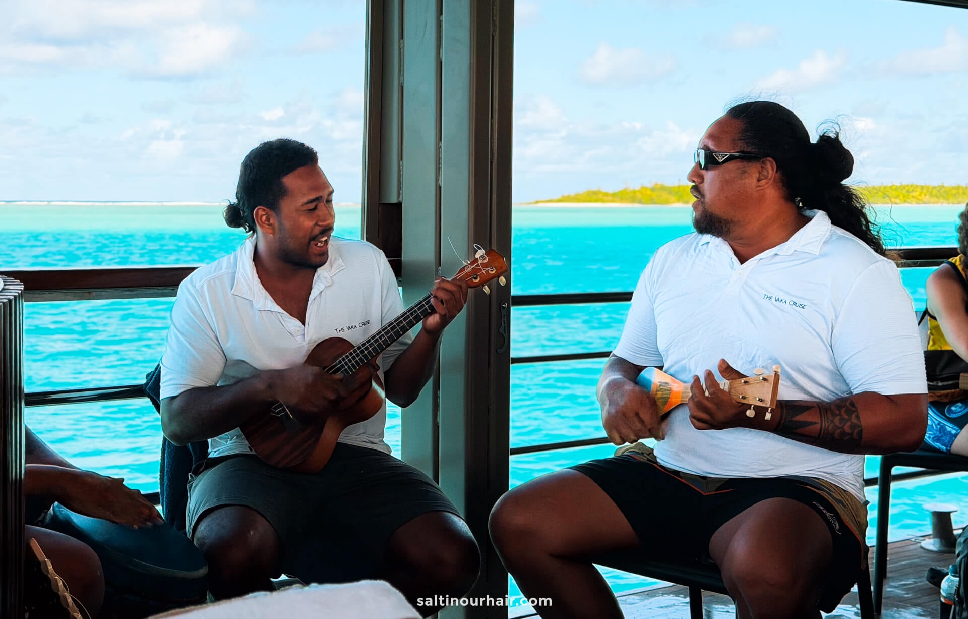 Two men in white shirts sit on a deck over turquoise water on Aitutaki island, playing ukuleles and singing, with an island and blue sky in the background.