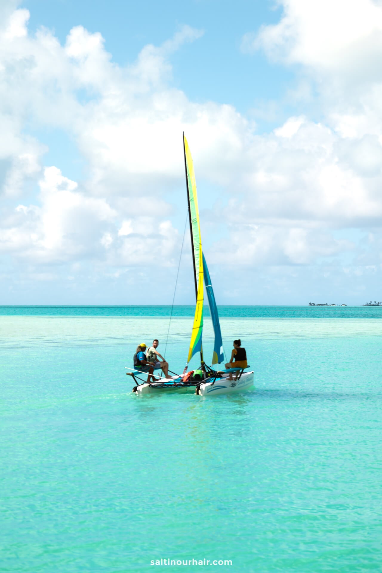 Three people sail a small catamaran with a blue and yellow sail on the clear turquoise waters of Aitutaki island, cook islands