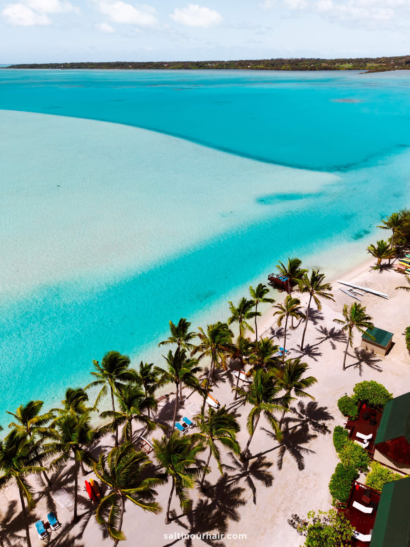Aerial view of Aitutaki island&rsquo;s tropical beach with clear turquoise water, palm trees lining the shore, and lounge chairs under umbrellas on the sandy coastline.