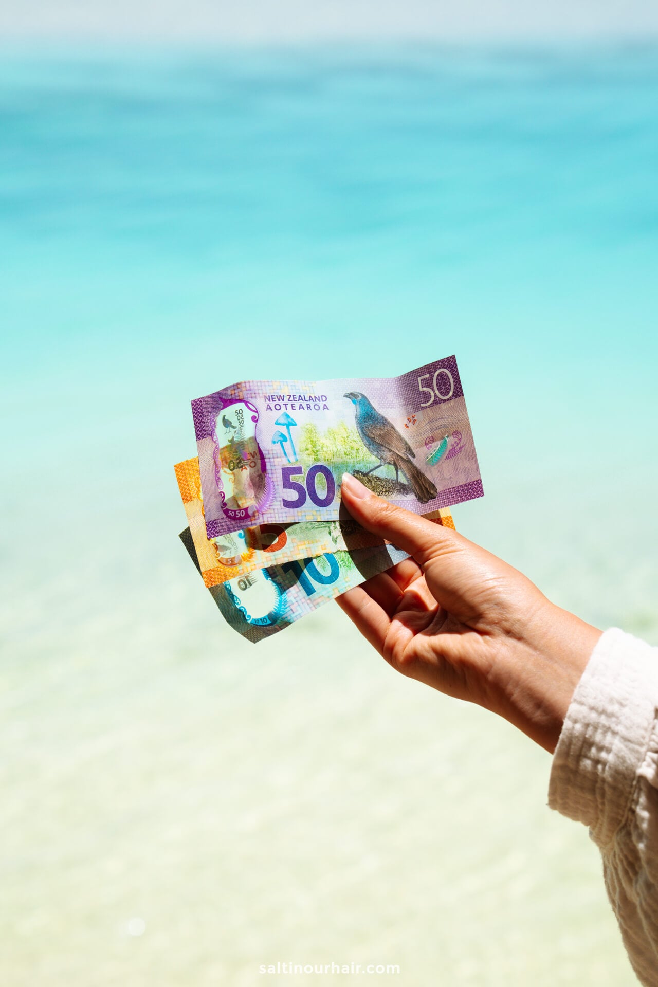 A hand holds New Zealand banknotes (50, 20, and 10 dollars) against the stunning blue ocean and sky of Aitutaki island in the background.