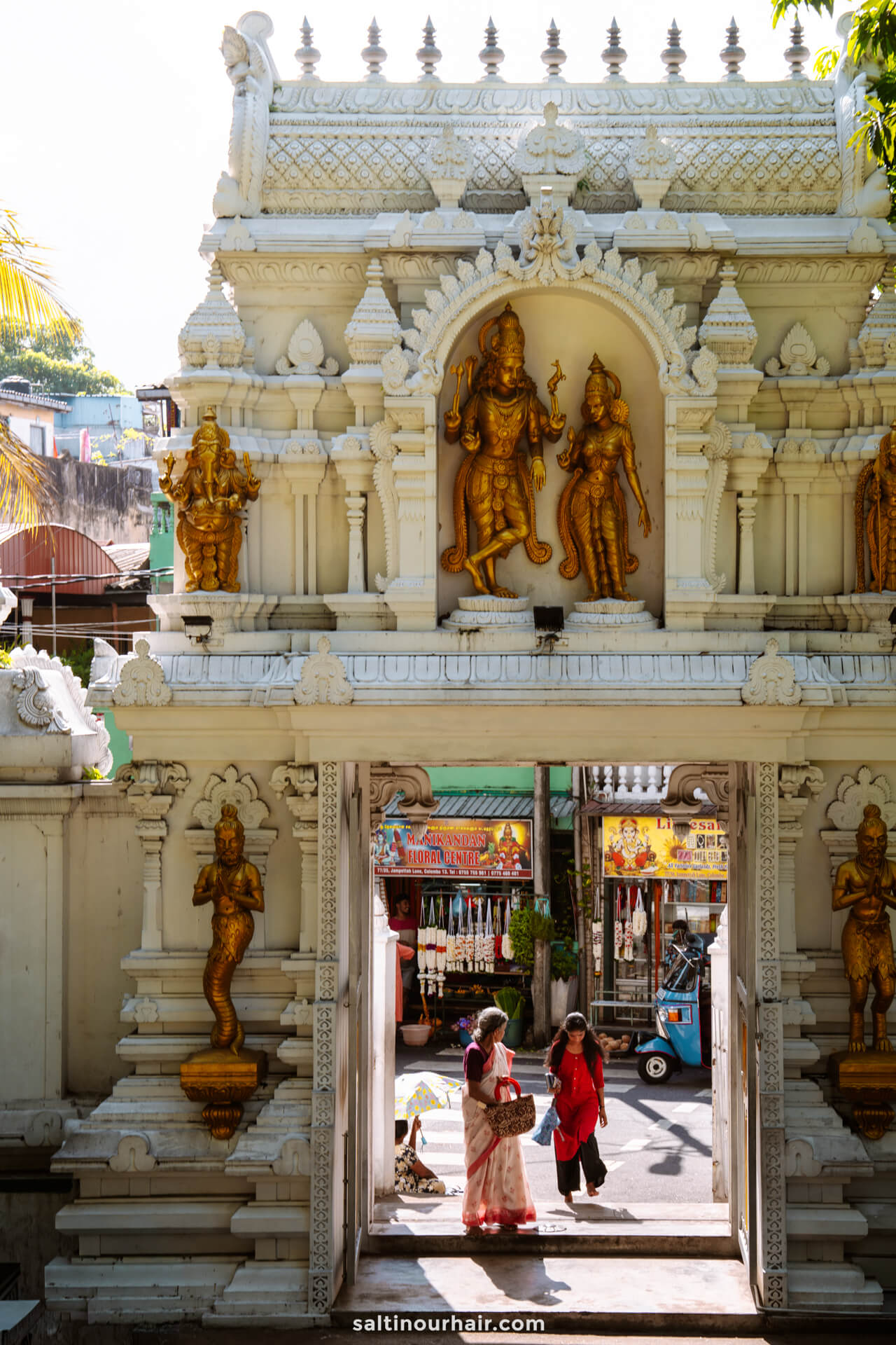 Ornate temple entrance with golden Hindu deities; two women in sarees stand at the open gate, with market stalls behind at Colombo, Sri Lanka.