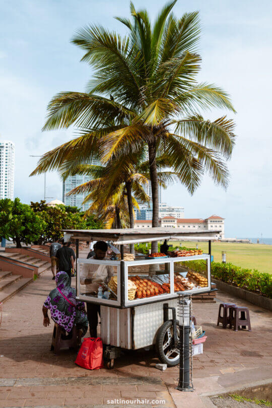 A street food cart displaying various snacks is parked under palm trees, with people sitting on stools nearby and city buildings at Galle Face Green-things to do in Colombo