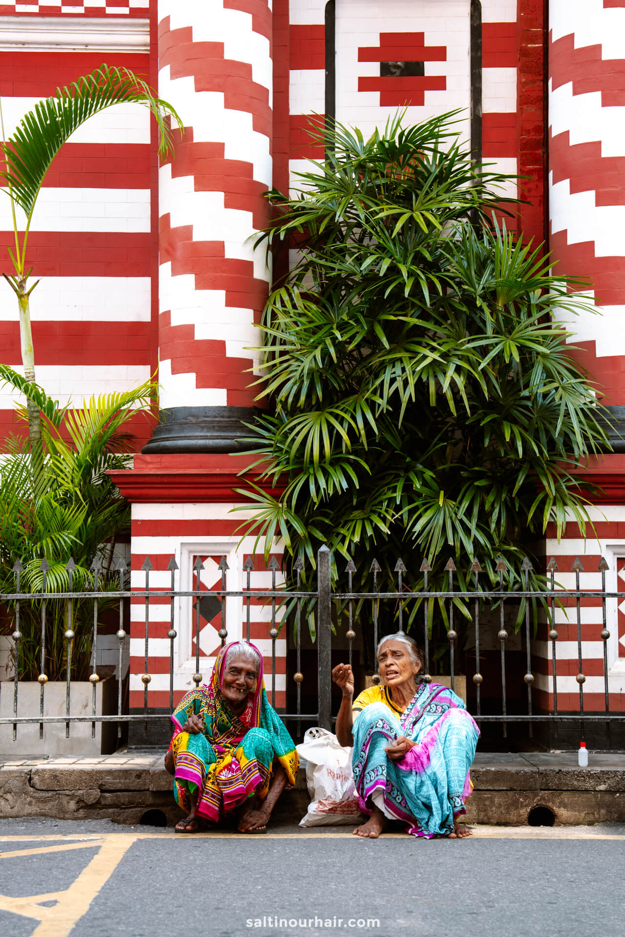 Two elderly women in colorful saris sit on the sidewalk in front of a red and white striped building with green plants&mdash;a vibrant scene that captures the charm found while exploring things to do Colombo.