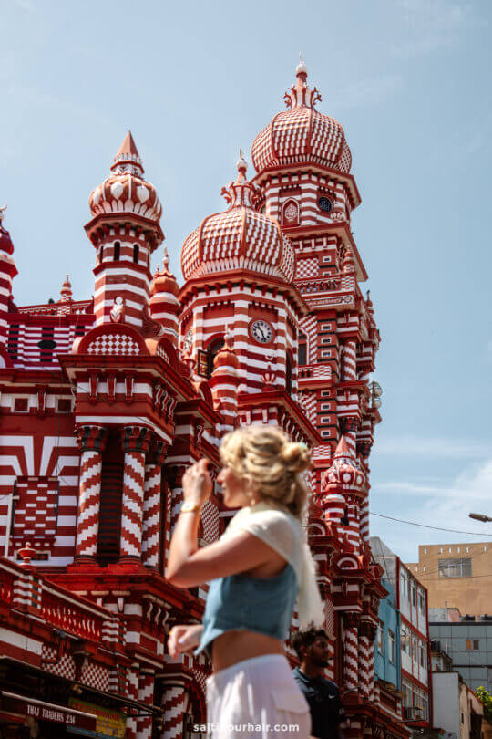 A woman walks past a red and white patterned mosque with domes and towers under a clear sky&mdash;a striking sight among the many things to do in Colombo.