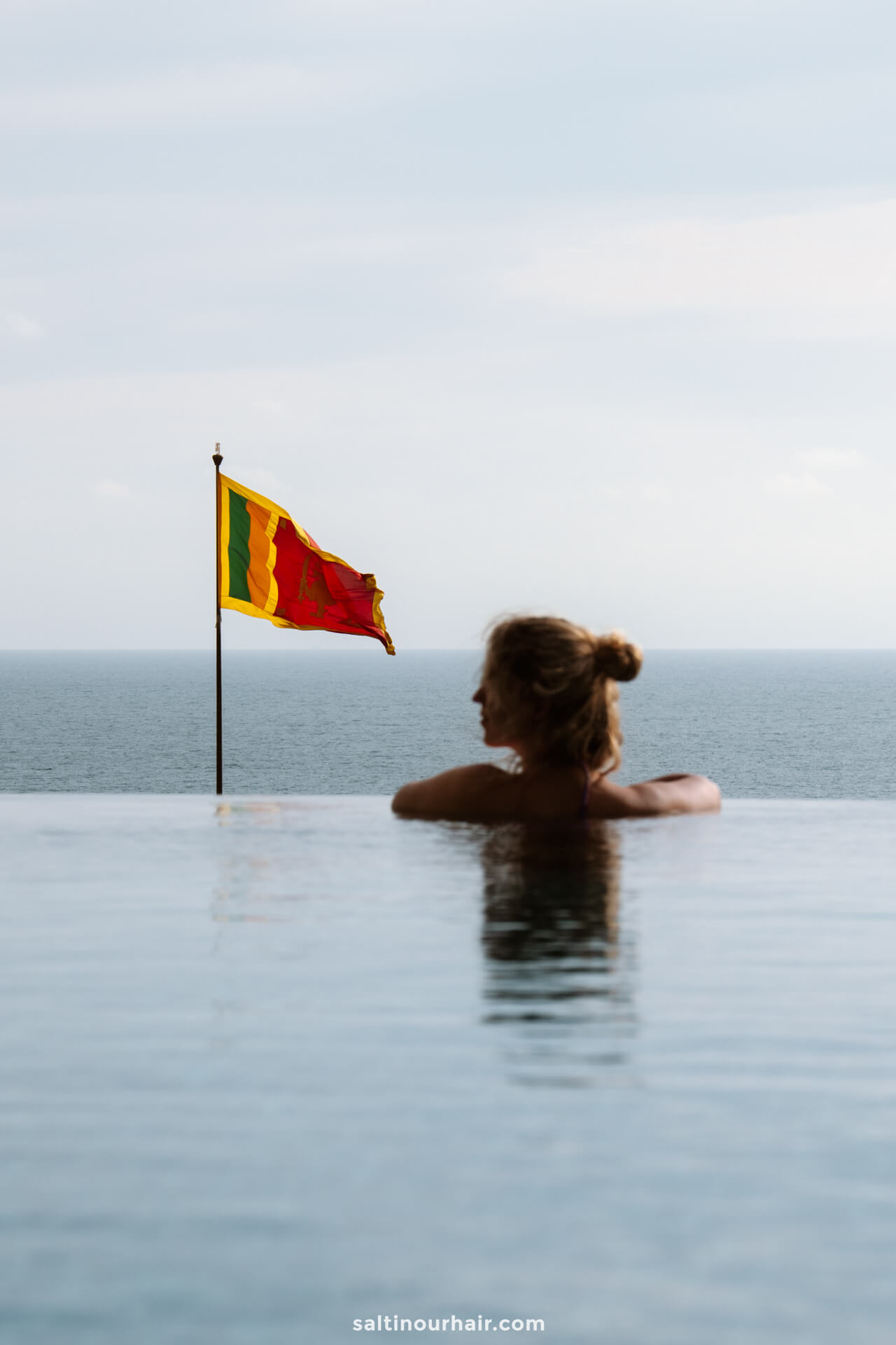 A person relaxes in an infinity pool overlooking the ocean, with a Sri Lankan flag visible in the background.