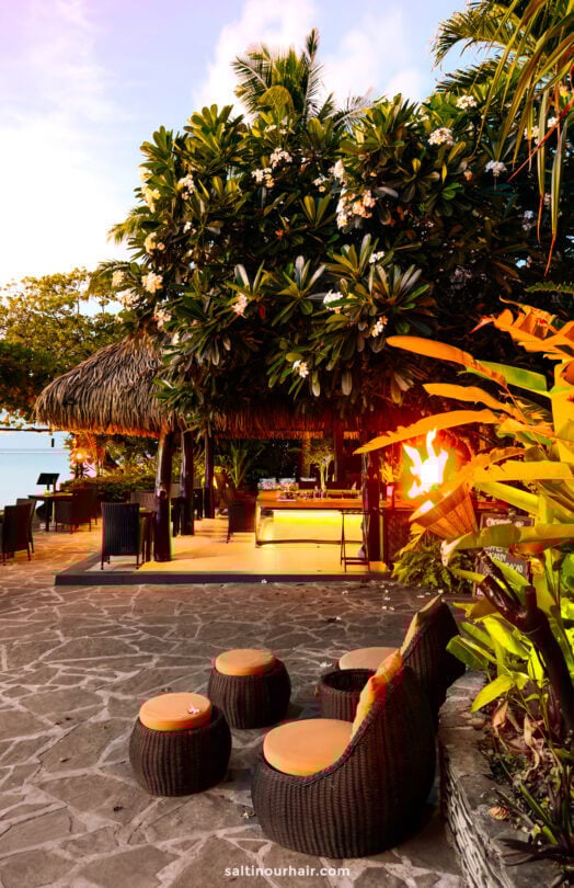 Outdoor tropical lounge area with wicker seating at pacific resort aitutaki, cook islands. a torch, and thatched-roof hut surrounded by lush greenery near the water at sunset