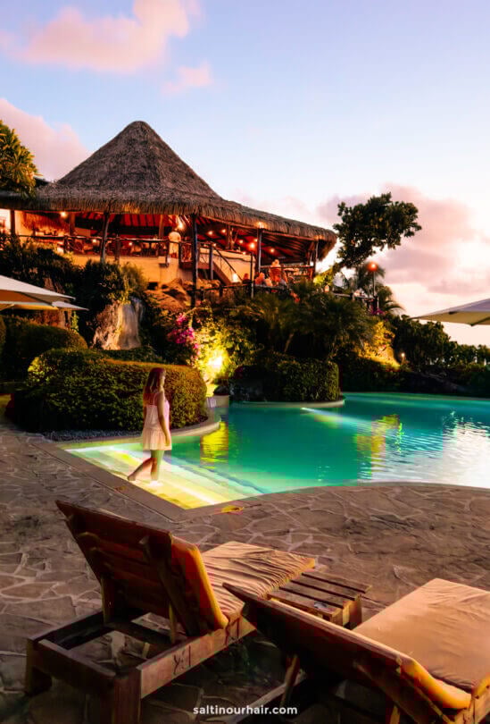 A woman in a white dress stands at the edge of a lit outdoor pool near a thatched-roof restaurant at dusk, at pacific beach resort Aitutaki Island, cook islands.
