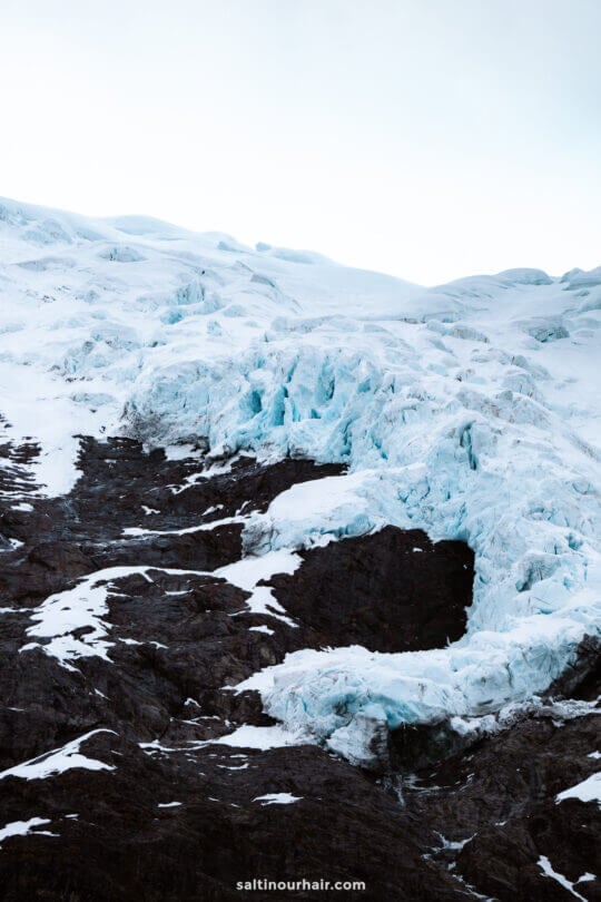 A glacier with blue and white ice sits atop a dark, rocky mountain slope under a pale sky along the scenic Rob Roy Track in New Zealand,