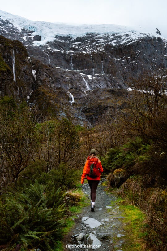 A person wearing a red jacket hikes on the rocky Rob Roy Track, surrounded by lush greenery, with a mountain and a glacier visible in the background.