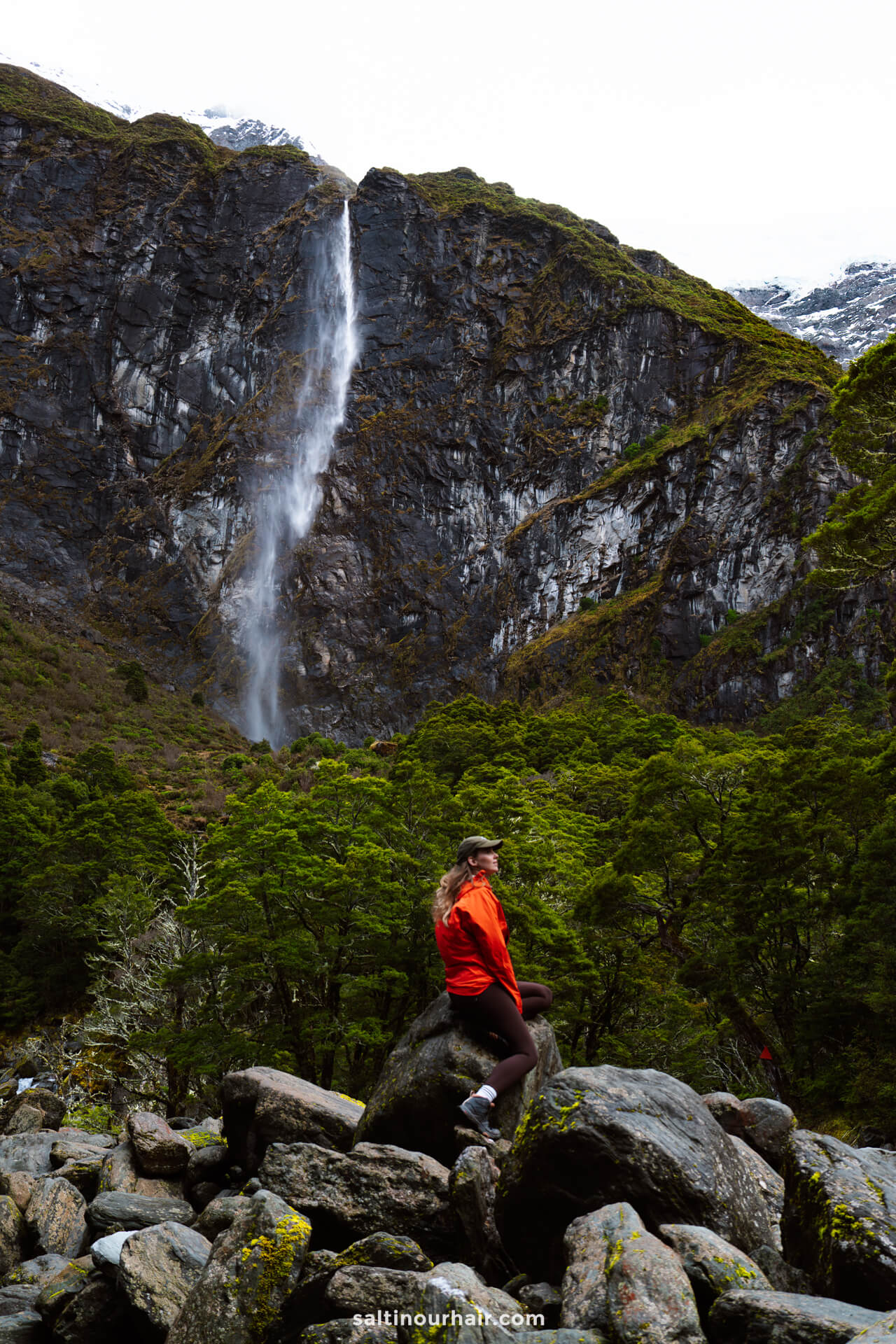 A woman in a red jacket sits on rocks along the Rob Roy Track in New Zealand, in front of a tall, narrow waterfall cascading down a steep, green mountainside under an overcast sky.