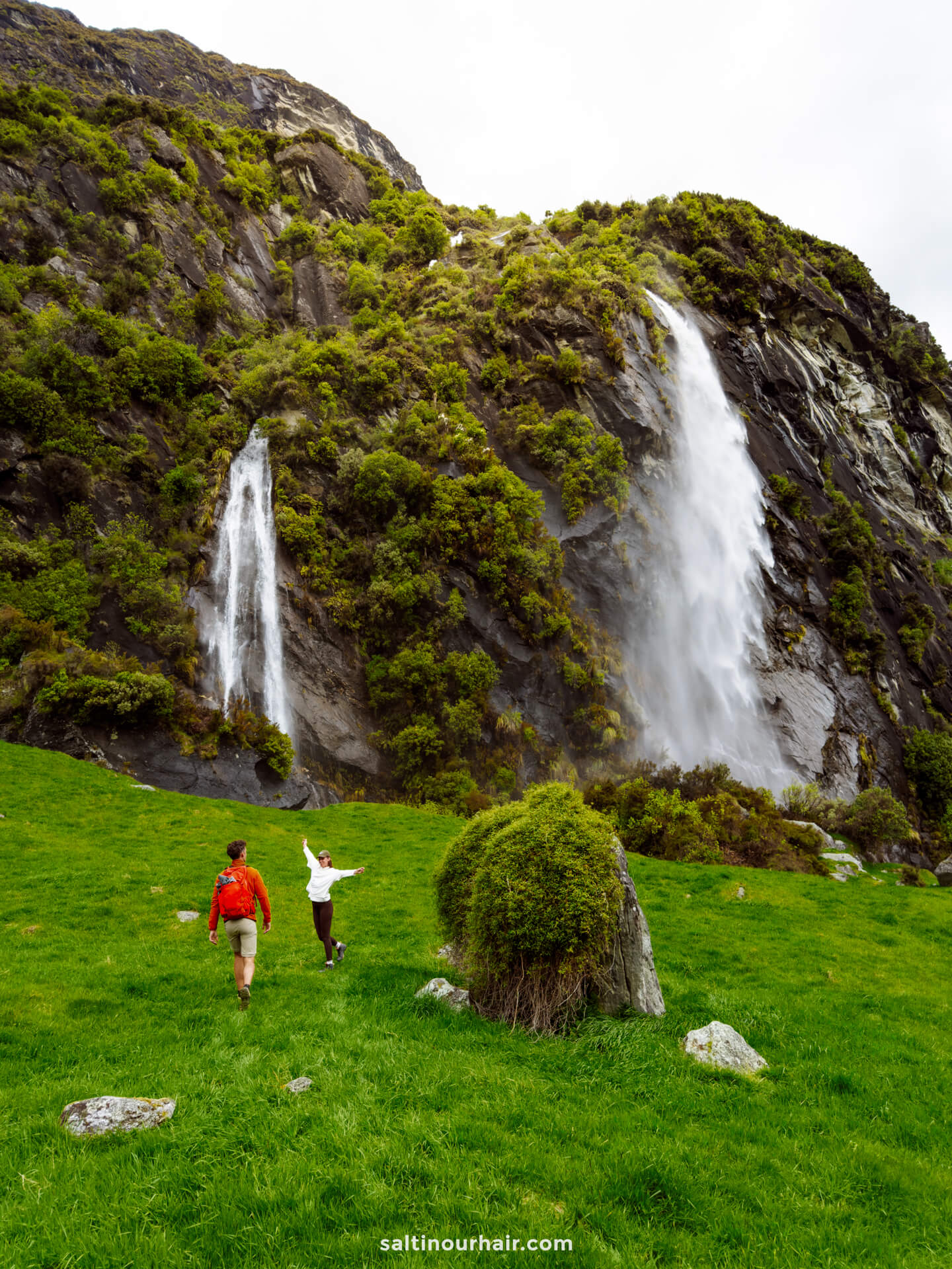 Two people walk on green grass toward a rocky hill with two waterfalls cascading down, surrounded by lush vegetation along the scenic Rob Roy Track in New Zealand,