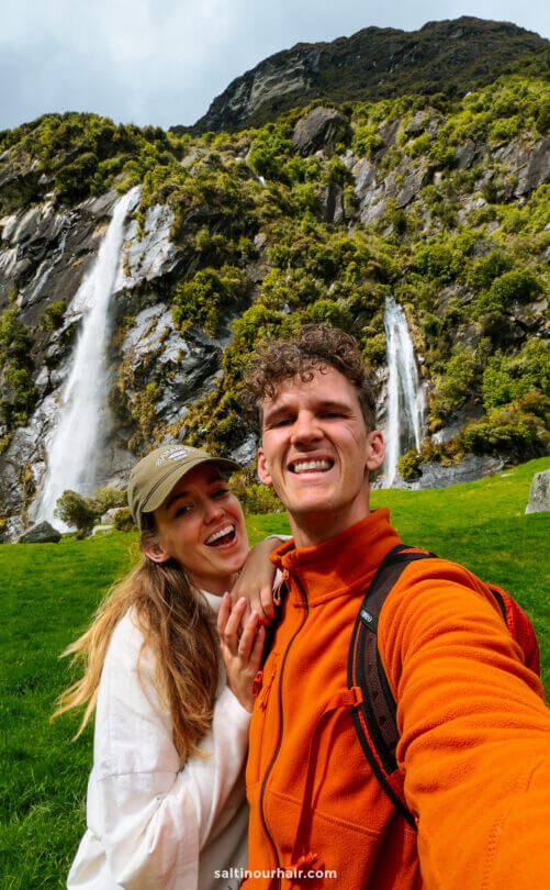 A man and woman smiling for a selfie in front of a grassy area with two waterfalls and rocky hills on the scenic Rob Roy Track in New Zealand,
