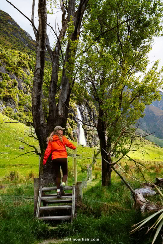 A woman in an orange jacket stands on a wooden platform along the Rob Roy Track, surrounded by lush green landscape, trees, and a majestic waterfall in the background.