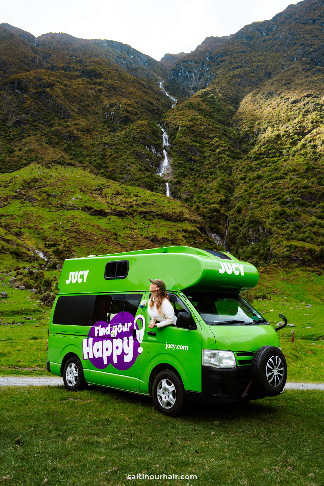 A woman in a hat leans out of the window of a bright green camper van parked on grass along the scenic Rob Roy Track in New Zealand,, with mountains and a waterfall in the background.
