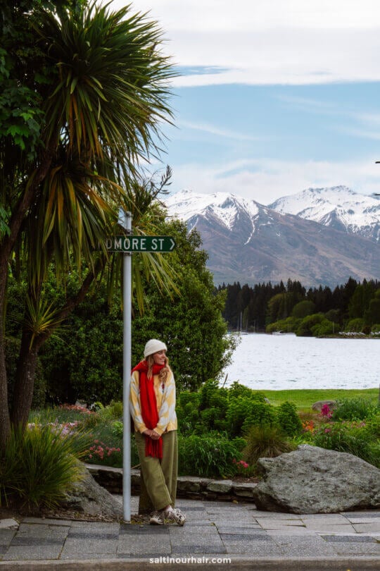 woman standing in wanaka new zealand wearing red scarf and white beenie and green plants, wanaka lake and snow capped mountains in the background