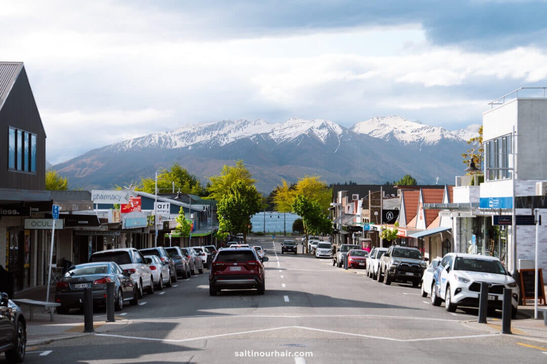 A quiet street in wanaka, new zealand, lined with parked cars and shops, with snow-capped mountains&mdash;reminiscent of those along the Rob Roy Track&mdash;and cloudy skies in the background.