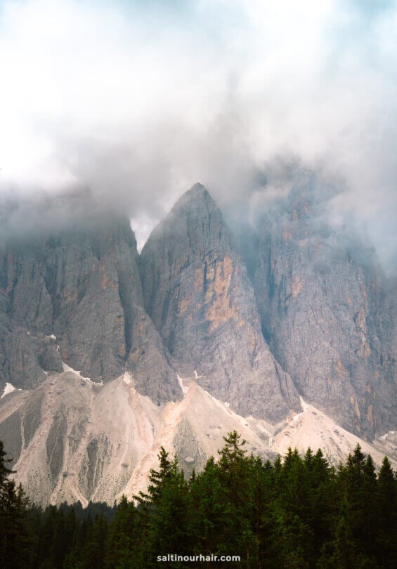 Jagged mountain peaks partly covered by clouds, with a forest of evergreen trees at the base&mdash;an iconic view along the Geisler Alm hike.
