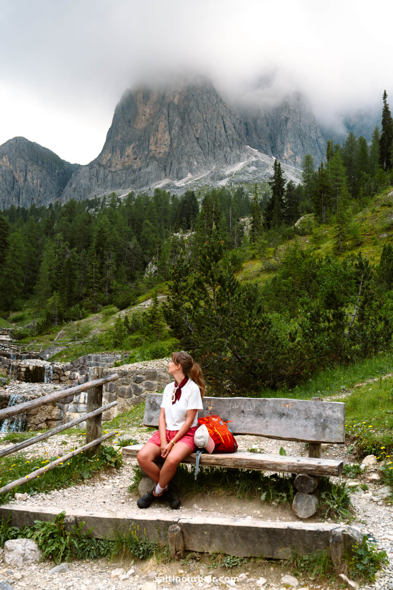 A person sits on a wooden bench with a red backpack, surrounded by trees and the stunning mountains of the Adolf Munkel Trail in the Dolomites, partly covered by clouds in the background.