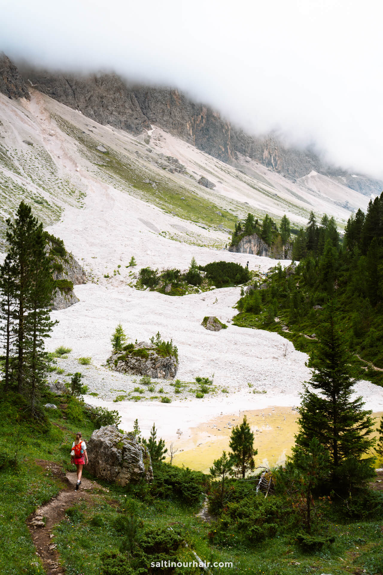 A hiker in a red jacket walks along the lush Adolf Munkel Trail to Geisler alm hut surrounded by rocky slopes, scattered trees, and low clouds drifting over the scenic mountain landscape.