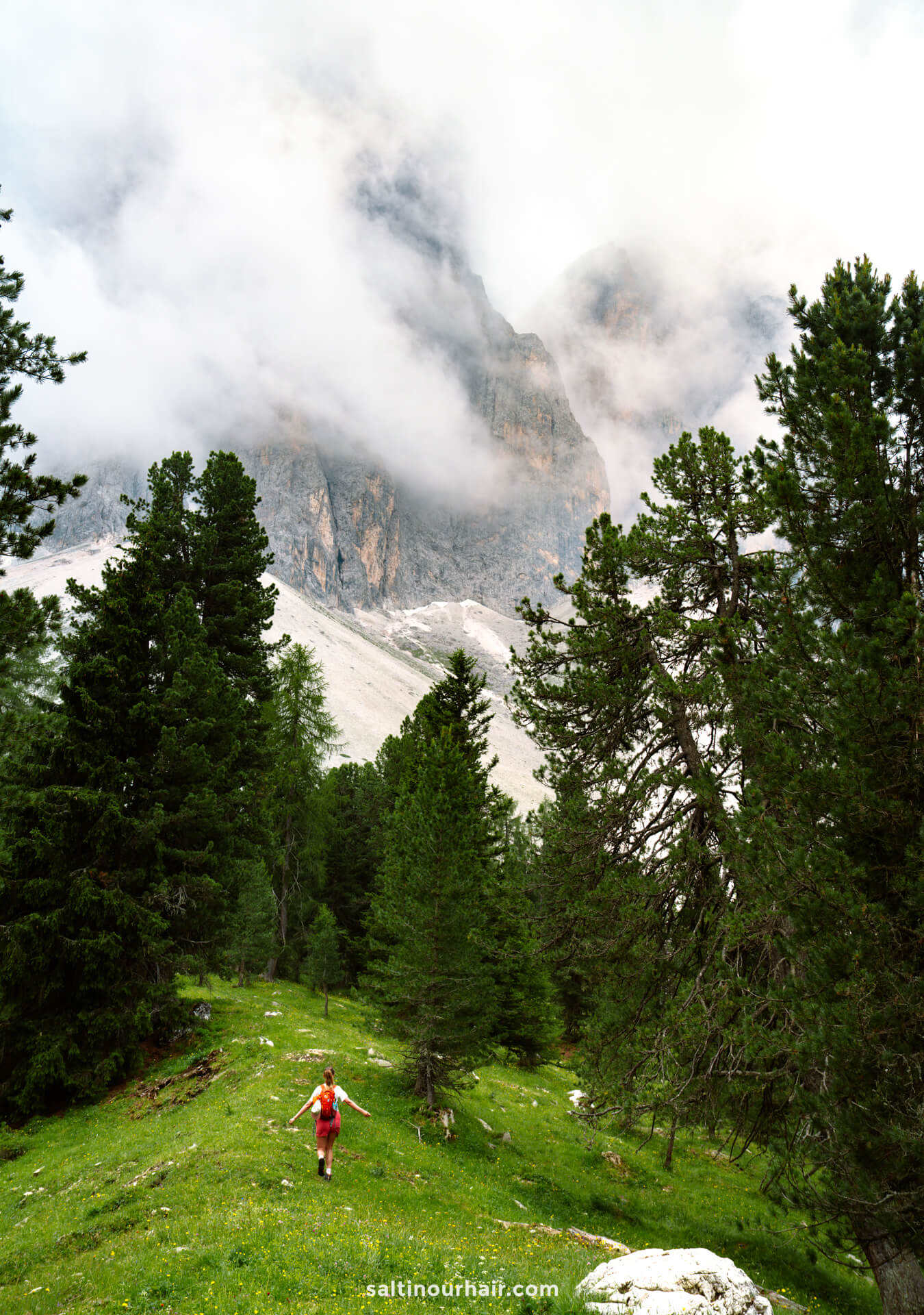 A person in a red outfit walks on a grassy slope surrounded by pine trees, with misty mountains and clouds in the background&mdash;capturing the serene beauty of the Geisler Alm hike in the Dolomites.