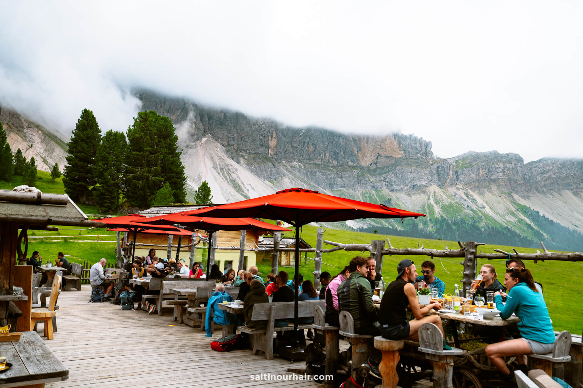 People dining outdoors at wooden tables under red umbrellas, with a scenic mountain landscape along the Adolf Munkel trail and a cloudy sky in the background.