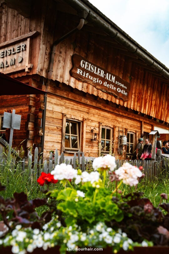 A rustic wooden mountain hut named Geisler-Alm sits behind a small garden with colorful flowers; people are gathered near its entrance after enjoying the scenic Geisler Alm hike.