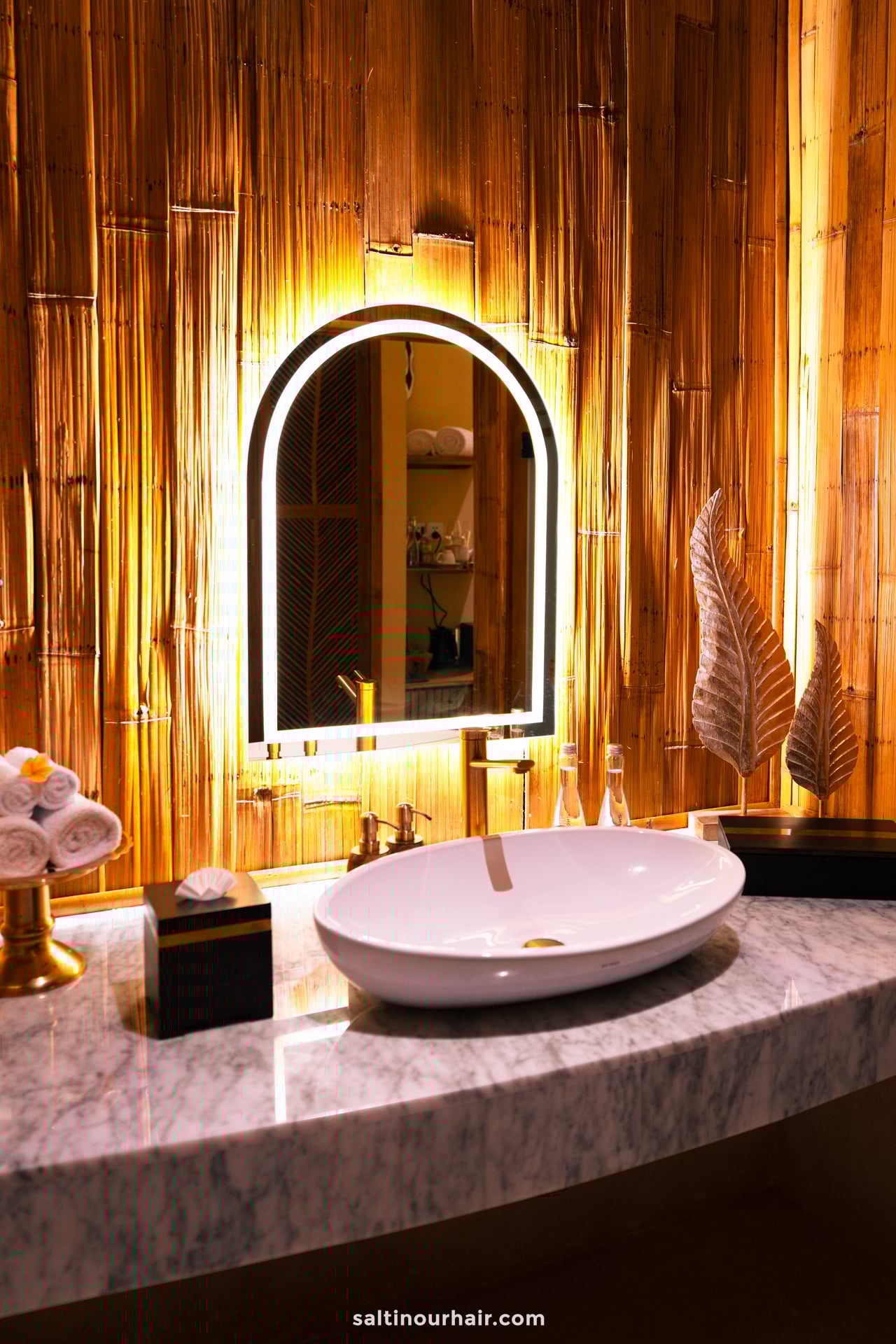 A modern bathroom vanity with a marble countertop, oval sink, backlit arched mirror, and decorative golden leaves creates a serene bambootel luxury hotel bali vibe against the bamboo hotel