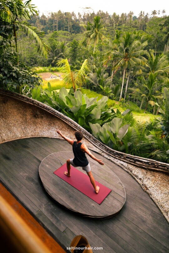 A person practices yoga on a red mat on a wooden deck at bambootel luxury hotel bali, surrounded by lush tropical greenery with a forest view in the background.