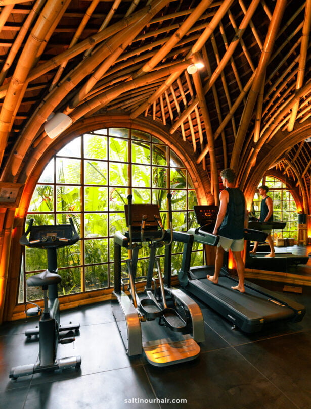 A person uses a treadmill in the bambootel sawah view luxury bamboo hotel bali gym with large arched windows and a bamboo ceiling, overlooking lush greenery outside.