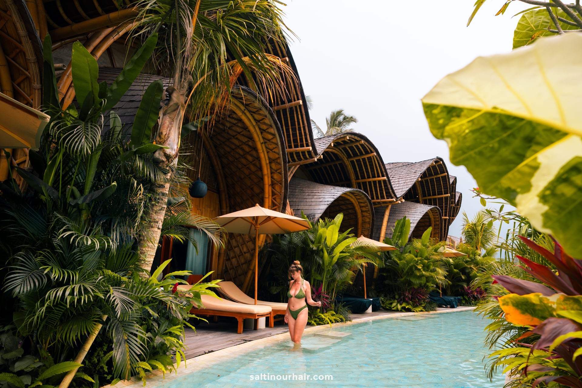 A woman in a green bikini walks by a pool surrounded by tropical plants and bambootel bamboo hotel villas in Bali with elegant, curved roofs.