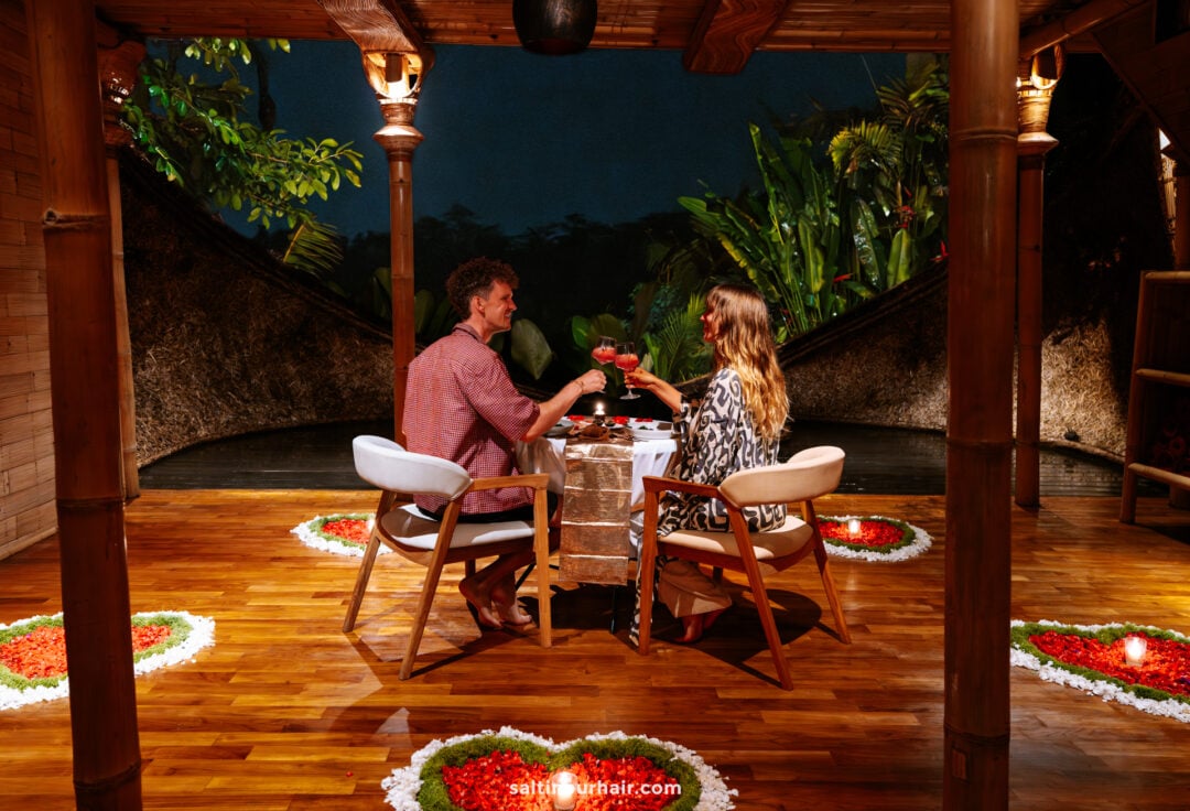A man and woman sit at a small table in a decorated wooden bambootel luxury hotel bali pavilion, toasting drinks, surrounded by floral arrangements, with greenery visible outside.
