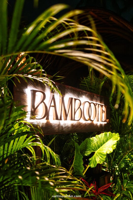 A wooden sign reading BAMBOOTEL illuminated among green tropical plants at night evokes the tranquil vibe of bambootel sawah view bali