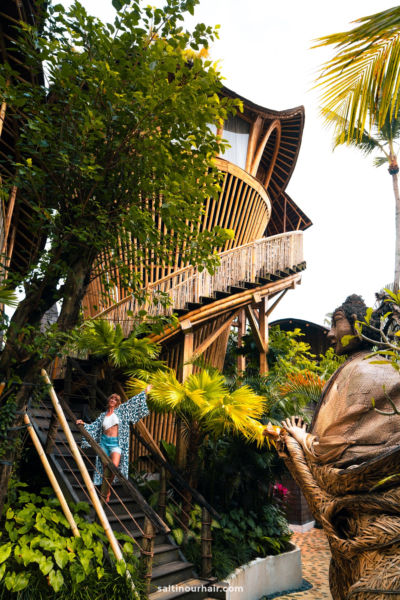 A woman stands on stairs leading to a large, modern bambootel bamboo hotel treehouse surrounded by tropical plants and trees in bali