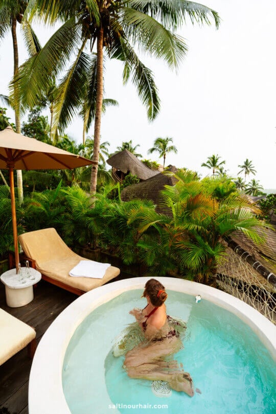 A woman with a flower in her hair relaxes in a round outdoor hot tub at a bambootel, surrounded by lush tropical plants and palm trees.