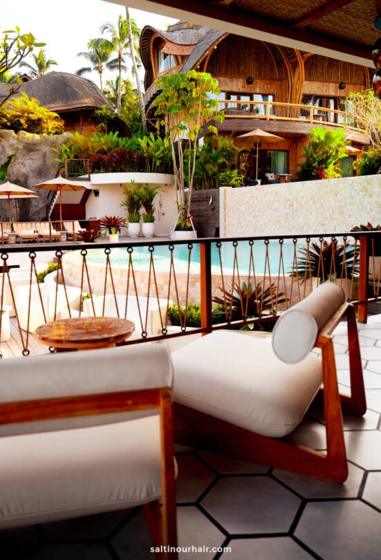 Outdoor seating area overlooking a pool, surrounded by tropical plants and a wooden bambootel luxury resort bali building in the background.