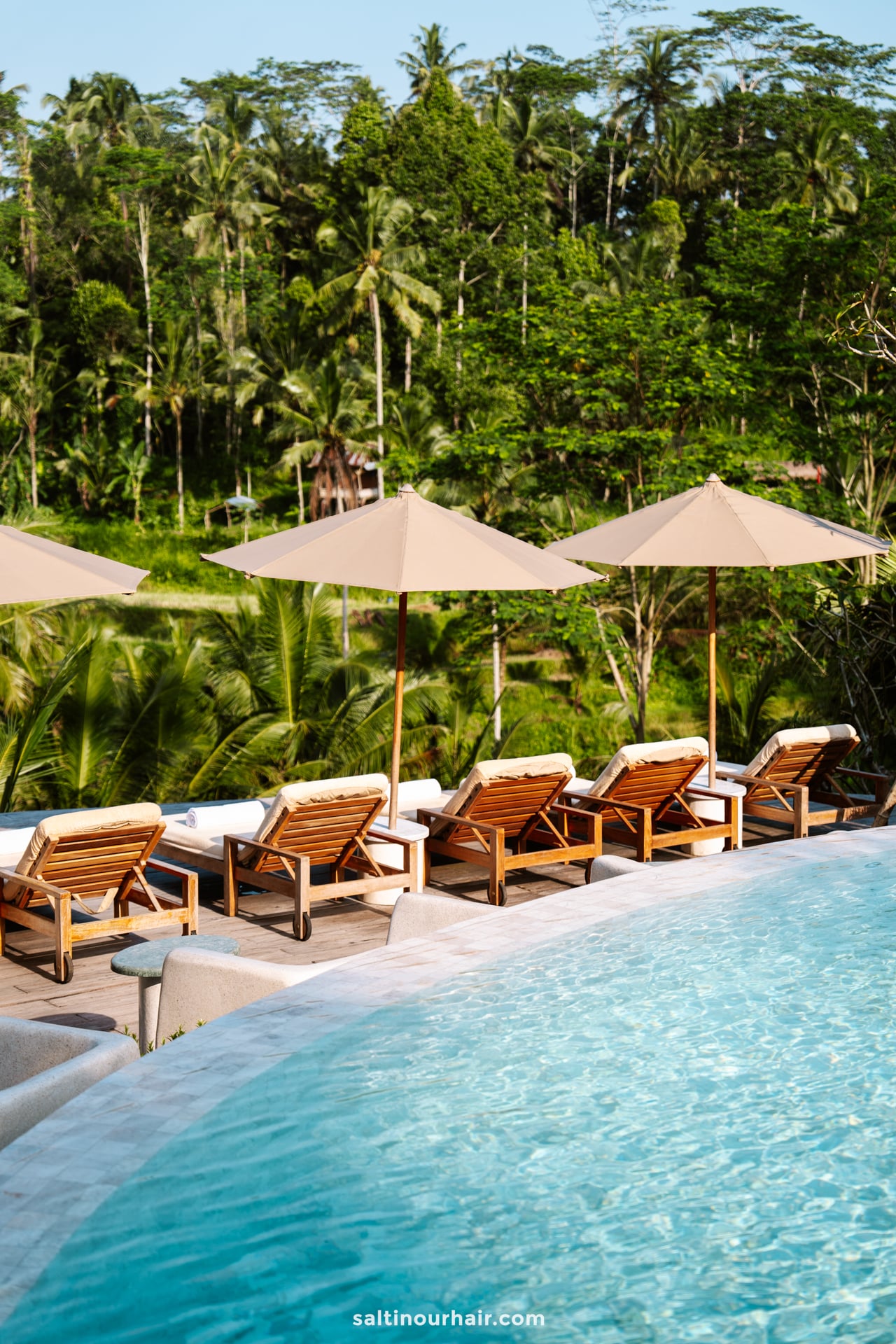 Wooden lounge chairs with beige umbrellas are lined up beside a curved pool at Bambootel resort bali overlooking lush green trees and tropical vegetation.