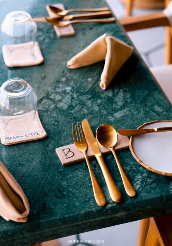 A green marble table at Bambootel sawah view bali is set with gold utensils, two upside-down glasses on coasters, and folded tan napkins.