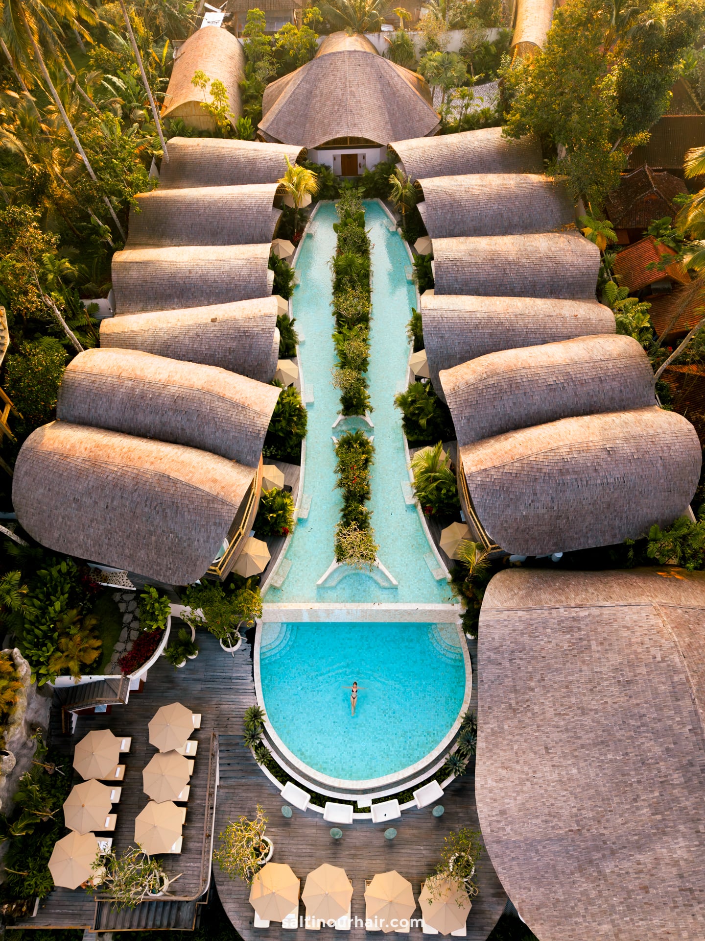 Aerial view of a bambootel bamboo hotel with thatched-roof villas surrounding a long, narrow pool leading to a round pool where a person is swimming; lounge chairs and umbrellas are nearby.