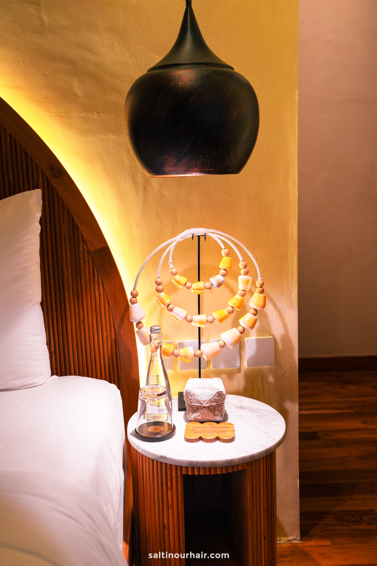 A bedside table at Bambootel holds a glass water bottle, a wrapped item, and a decorative wooden bead ornament, set next to the bed and beneath a black pendant light. luxury eco hotel bali
